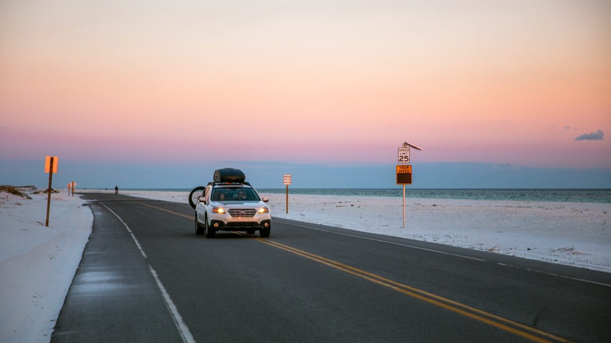 A car drives along a beach road lined with white sand as the sky turns pink at sunset over the Gulf.
