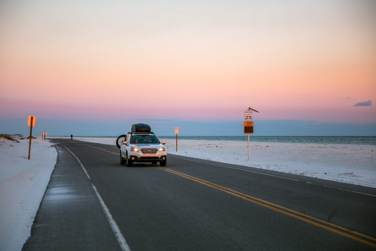 A car drives along a beach road lined with white sand as the sky turns pink at sunset over the Gulf.