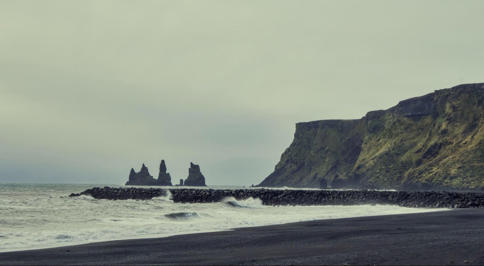 Waves on the black sand beach of Reynisfjara, with sea stacks offshore and a mossy cliff under an overcast sky
