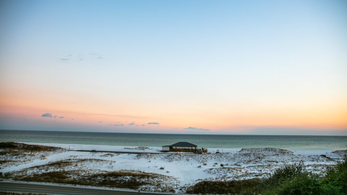 A beach pavilion sits among white dunes beside the Gulf of Mexico under a pale pink sunrise sky.