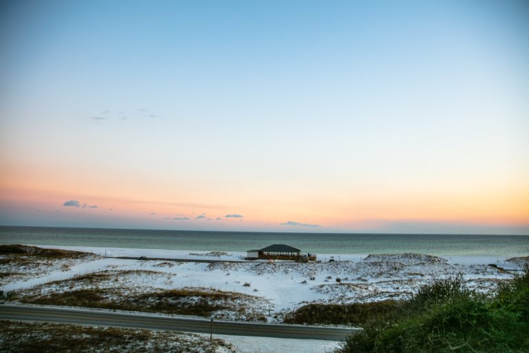A beach pavilion sits among white dunes beside the Gulf of Mexico under a pale pink sunrise sky.
